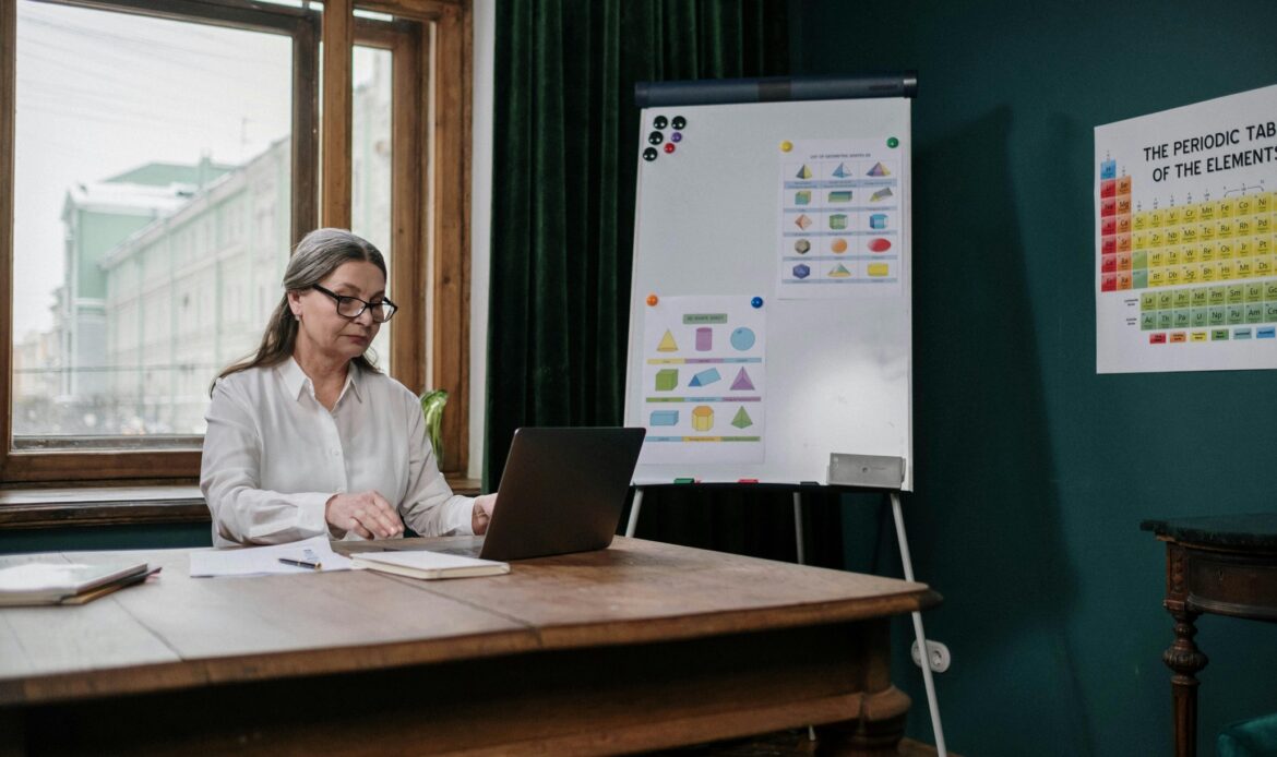 Female professor using a laptop while seated at a table in a classroom